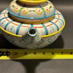 Puede incluir: Una tetera de cer&aacute;mica blanca con tapa, con un dise&ntilde;o pintado a mano con detalles en azul, naranja y amarillo. La tetera tiene un pico y un asa. La tetera mide aproximadamente 15 cm de di&aacute;metro.