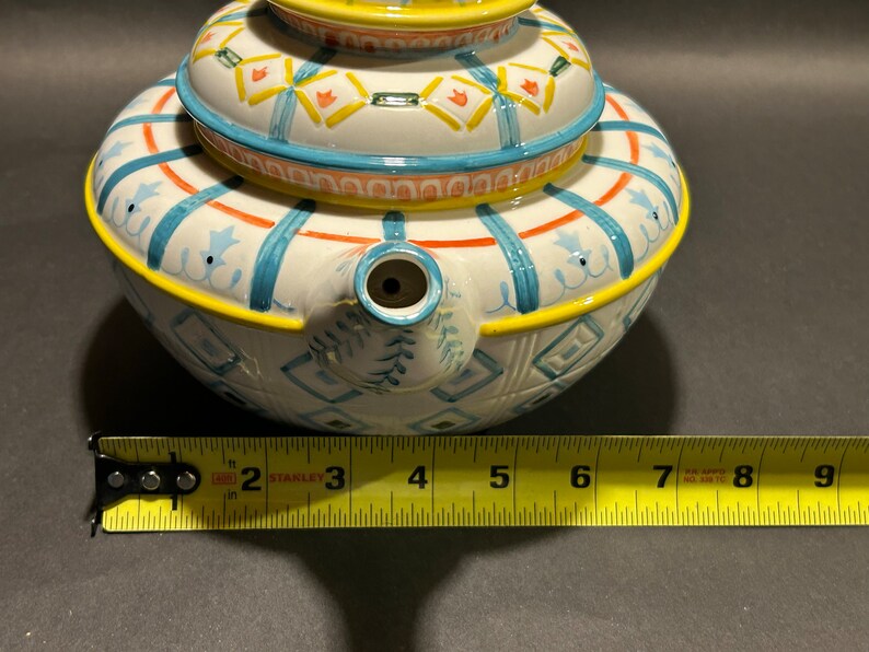 Puede incluir: Una tetera de cer&aacute;mica blanca con tapa, con un dise&ntilde;o pintado a mano con detalles en azul, naranja y amarillo. La tetera tiene un pico y un asa. La tetera mide aproximadamente 15 cm de di&aacute;metro.