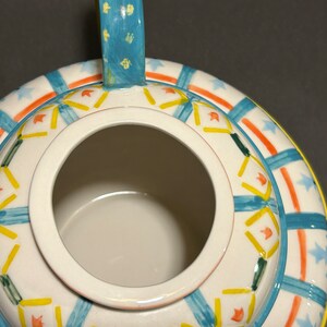 Puede incluir: Vista superior de una tetera de cer&aacute;mica blanca con asa y borde azules. La tetera est&aacute; decorada con patrones geom&eacute;tricos y formas de estrella azules, naranjas, amarillas y verdes. El interior de la tetera es blanco.
