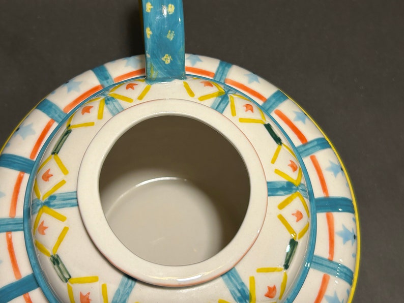 Puede incluir: Vista superior de una tetera de cer&aacute;mica blanca con asa y borde azules. La tetera est&aacute; decorada con patrones geom&eacute;tricos y formas de estrella azules, naranjas, amarillas y verdes. El interior de la tetera es blanco.
