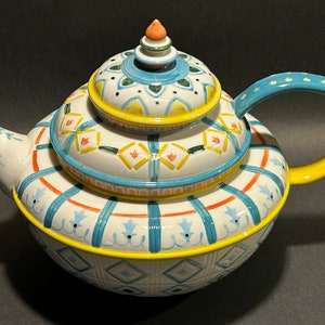 Puede incluir: Una tetera de cer&aacute;mica decorativa con tapa a juego. La tetera es blanca con patrones geom&eacute;tricos azules, naranjas y amarillos. El asa es amarilla y la tapa tiene un peque&ntilde;o adorno naranja. La tetera est&aacute; dise&ntilde;ada para servir t&eacute;.