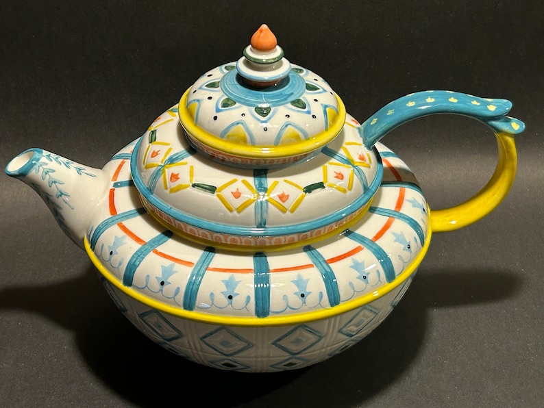 Puede incluir: Una tetera de cer&aacute;mica decorativa con tapa a juego. La tetera es blanca con patrones geom&eacute;tricos azules, naranjas y amarillos. El asa es amarilla y la tapa tiene un peque&ntilde;o adorno naranja. La tetera est&aacute; dise&ntilde;ada para servir t&eacute;.