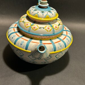 Puede incluir: Una tetera de cer&aacute;mica blanca con tapa, con un dise&ntilde;o apilado. La tetera est&aacute; adornada con patrones geom&eacute;tricos azules, amarillos y naranjas. La boquilla es visible en el lateral. El asa y el pomo de la tapa tambi&eacute;n est&aacute;n decorados.