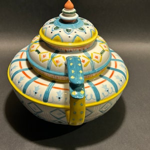Puede incluir: Una tetera de cer&aacute;mica decorativa con un dise&ntilde;o colorido. La tetera presenta una base blanca con detalles en azul, naranja y amarillo. La tapa y el asa tambi&eacute;n est&aacute;n decoradas con patrones intrincados. La tetera tiene una forma redondeada y un peque&ntilde;o adorno decorativo en la parte superior.