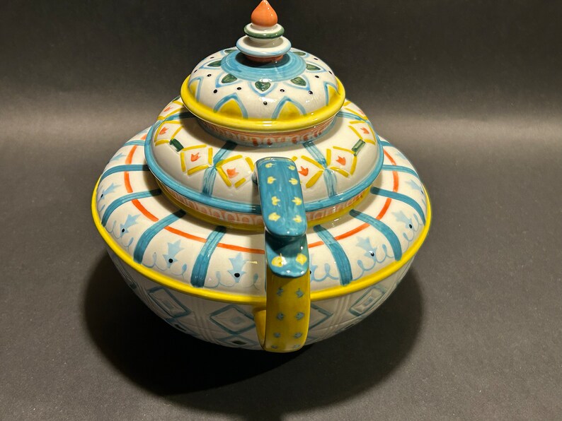 Puede incluir: Una tetera de cer&aacute;mica decorativa con un dise&ntilde;o colorido. La tetera presenta una base blanca con detalles en azul, naranja y amarillo. La tapa y el asa tambi&eacute;n est&aacute;n decoradas con patrones intrincados. La tetera tiene una forma redondeada y un peque&ntilde;o adorno decorativo en la parte superior.