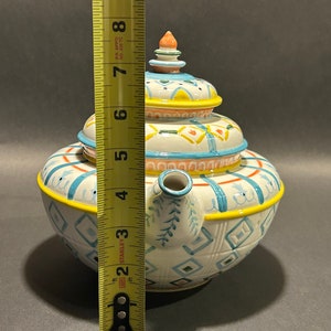 Puede incluir: Una tetera de cer&aacute;mica decorativa con tapa, de aproximadamente 18 cm de altura. La tetera es blanca con patrones geom&eacute;tricos azules, amarillos y naranjas. Tiene un pico y un asa, y est&aacute; dise&ntilde;ada para servir t&eacute;.
