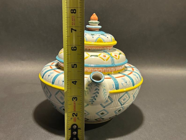 Puede incluir: Una tetera de cer&aacute;mica decorativa con tapa, de aproximadamente 18 cm de altura. La tetera es blanca con patrones geom&eacute;tricos azules, amarillos y naranjas. Tiene un pico y un asa, y est&aacute; dise&ntilde;ada para servir t&eacute;.