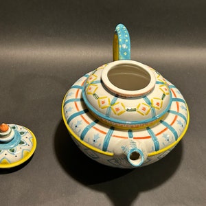 Puede incluir: Una tetera de cer&aacute;mica decorativa con tapa a juego. La tetera es blanca con patrones geom&eacute;tricos azules, amarillos y naranjas. El asa es azul claro con lunares amarillos. La tapa est&aacute; al lado de la tetera.