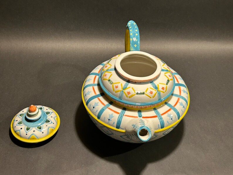 Puede incluir: Una tetera de cer&aacute;mica decorativa con tapa a juego. La tetera es blanca con patrones geom&eacute;tricos azules, amarillos y naranjas. El asa es azul claro con lunares amarillos. La tapa est&aacute; al lado de la tetera.