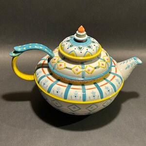 Puede incluir: Una tetera de cer&aacute;mica decorativa con tapa. La tetera presenta una base blanca con patrones geom&eacute;tricos azules, amarillos y naranjas. El asa es amarilla con detalles azules. La tapa est&aacute; rematada con un peque&ntilde;o adorno naranja. La tetera est&aacute; dise&ntilde;ada para servir t&eacute;.