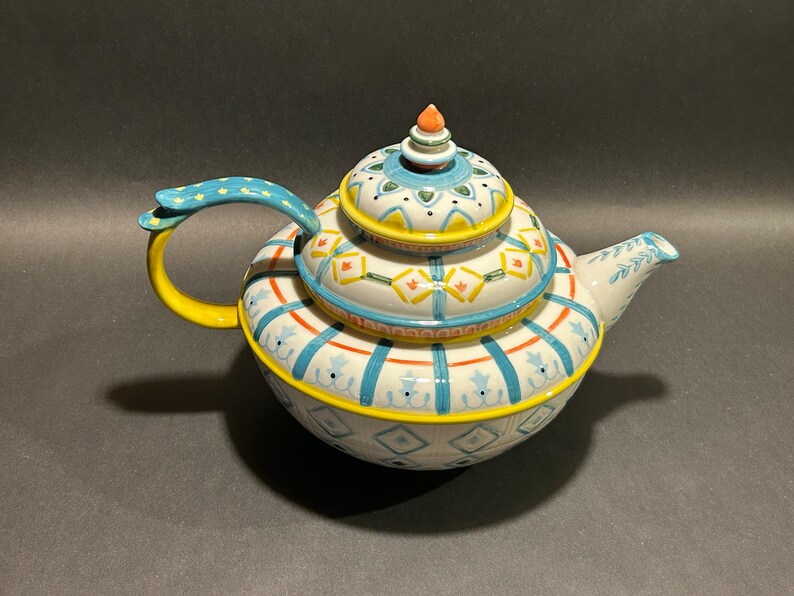 Puede incluir: Una tetera de cer&aacute;mica decorativa con tapa. La tetera presenta una base blanca con patrones geom&eacute;tricos azules, amarillos y naranjas. El asa es amarilla con detalles azules. La tapa est&aacute; rematada con un peque&ntilde;o adorno naranja. La tetera est&aacute; dise&ntilde;ada para servir t&eacute;.