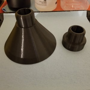 May include: Two black plastic 3D printed funnels of different sizes. The larger funnel is cone-shaped with a wide opening and a smaller opening at the top. The smaller funnel is cylindrical with a smaller opening at the top.