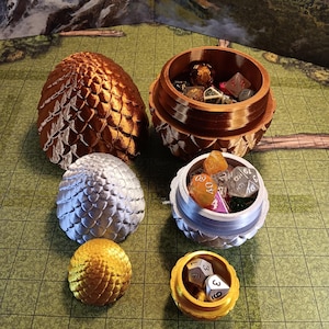 May include: A collection of dragon egg-shaped dice containers in various colors, including gold, silver, and bronze. The containers are open, revealing dice inside. The containers are on a green surface.