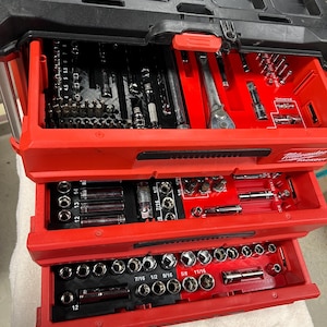 以下が含まれることがあります： 赤と黒のMilwaukee Packoutツールボックス。3つの引き出しが開いています。引き出しには、ソケット、ビット、レンチなど、さまざまなツールが収納されています。Milwaukee Packoutのロゴが上部と引き出しに表示されています。