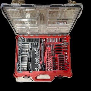 May include: Open red toolbox with a clear lid, filled with various metal tools. The tools are neatly organized in compartments. The toolbox has a handle and is set against a black background. The brand name "Milwaukee" is visible on the lid.