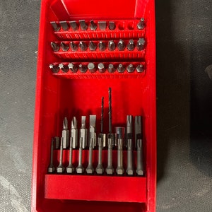 Könnte beinhalten: Ein roter Kunststoff-Aufbewahrungskasten mit einer Auswahl an Bohrer- und Schraubendreherbits. Der Kasten hat ein Fach zur Aufbewahrung der Bits und ein separates Fach zur Aufbewahrung eines Bohrers.