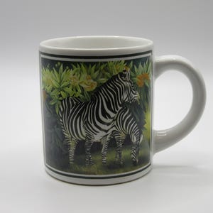 May include: A white ceramic mug featuring a rectangular graphic print of two zebras standing in a lush green environment with yellow flowers. The mug has a smooth finish, a curved handle on the right side, and a black border framing the zebra illustration.