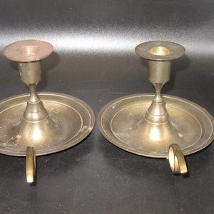 May include: A pair of matching brass candle holders, each featuring a wide circular base with a small side handle and a central stem topped by a flat circular candle cup. The metal has a muted, aged finish with visible signs of wear.