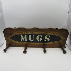 May include: A rectangular wooden wall rack with a dark brown finish, featuring four pegs for hanging items. The center displays an oval black plaque with the word MUGS in white capital letters, surrounded by a decorative gold-colored border.