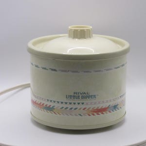 May include: A cream-colored Rival Little Dipper electric stoneware warmer with a matching lid. The cylindrical base features a decorative horizontal band with colorful geometric patterns and the text Rival Little Dipper Electric Stoneware Series.