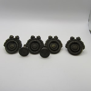 May include: A set of four dark bronze-toned metal drawer pulls featuring a circular design with a central floral motif and a swinging ring handle, accompanied by two smaller matching circular knobs with a similar central floral pattern.