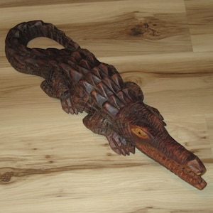May include: A dark brown wooden sculpture of a crocodile with a textured, scaled back and a curved tail. The figure features carved details along the body, four legs with visible claws, and an elongated snout with an open mouth showing teeth.