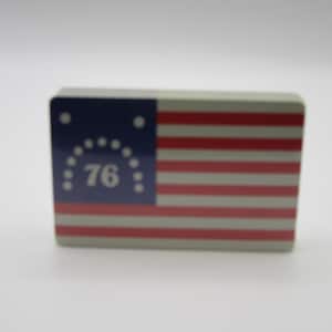 May include: A rectangular object featuring a graphic of the Bennington flag. The design includes a blue canton with a white number 76 surrounded by a circular arrangement of thirteen white stars, alongside horizontal red and white stripes.