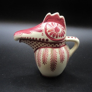 May include: A cream-colored ceramic creamer with a red rooster-shaped spout and handle. The creamer is decorated with red floral and geometric patterns. The design includes a detailed rooster head and a small handle for easy pouring.