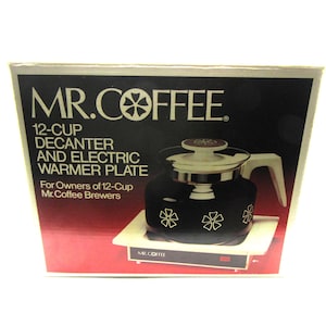 May include: A Mr. Coffee 12-cup decanter and electric warmer plate in its original packaging. The black glass decanter features a white floral pattern, a white lid, and a white handle. The white electric warmer plate has a small red power indicator.