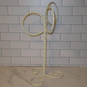 May include: A cream-colored metal stand featuring a central vertical twisted rod, a three-legged base with curled feet, and two circular twisted loops at the top designed for hanging items. The structure is made of twisted wire throughout.