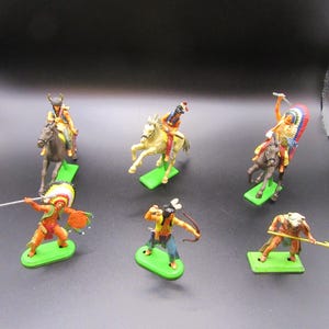 May include: A collection of vintage toy figurines depicting Native American figures, including warriors on horseback and standing figures with weapons. The figurines are painted in bright colors and mounted on green bases.