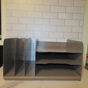 May include: A taupe metal desk organizer featuring three vertical slots on the left side for document storage and three horizontal stacked trays on the right side for paper organization, all integrated into a single solid rectangular base unit.