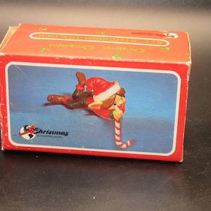 May include: A rectangular red cardboard box featuring a graphic of an elf wearing a red and white hat, lying down and holding a red and white striped candy cane. The box text reads Christmas Ornament and Elf Stocking Holder with a logo.