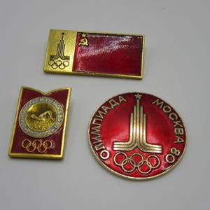May include: Three metal pins featuring red enamel and gold-tone accents. One rectangular pin displays the Soviet hammer and sickle. A second rectangular pin shows a swimmer. A circular pin features the Moscow 80 Olympic logo and text.