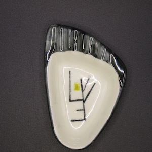 May include: A ceramic ashtray with a unique, foot-shaped design. The ashtray features a white interior with a black rim and a stylized black and yellow graphic. The overall aesthetic is modern and artistic.