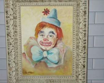 May include: An oil painting of a clown with orange hair, a light blue hat with a red flower, and a large light blue bow tie, wearing white face makeup with red lips and nose, housed in an ornate, distressed off-white rectangular frame.