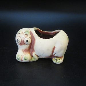 May include: A ceramic planter shaped like a hippopotamus with a cream-colored body, brown accents around the eyes and back, and large protruding eyes. The hollow opening is located on the back of the animal figure, which features a smooth finish.