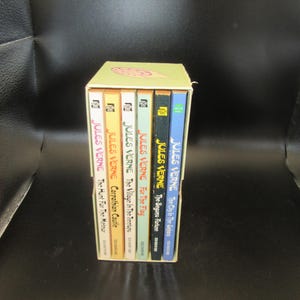 May include: A boxed set of Jules Verne books. The spines are visible, displaying titles like "The City in the Sahara" and "The Hunt For The Meteor." The box is light yellow with a floral design on top. The books have various colored spines.