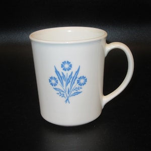 May include: A white ceramic mug featuring a blue floral pattern on the front. The design consists of three stylized flowers with pointed leaves and stems. The mug has a smooth, cylindrical body and a single curved handle on the right side.