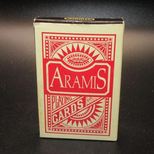 May include: A rectangular deck of Aramis playing cards in a cream-colored box with red geometric patterns and borders. The front of the box features the word Aramis in a stylized font above the words Playing Cards, all printed in red ink.