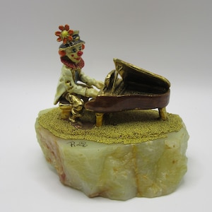 May include: A metal figurine of a clown wearing a hat with an orange flower, seated at a brown grand piano. The figure is mounted on a textured, light green stone base with gold-colored granular accents and the signature Piron inscribed on the front.