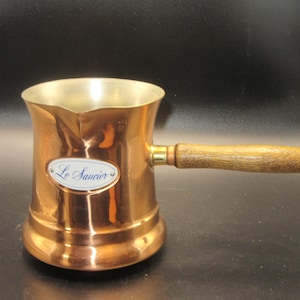 May include: A copper-colored metal sauce pot with a pouring spout and a straight wooden handle. An oval white label on the front features the blue text Le Saucier. The interior of the pot is a metallic silver color, and the exterior is polished.