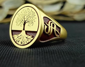Puede incluir: Un anillo de oro con una banda de esmalte rojo y un diseño de árbol de la vida en la parte delantera. El árbol está grabado en detalle y tiene un tronco grueso y muchas ramas.