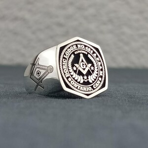 Puede incluir: Anillo mas&oacute;nico de plata con una incrustaci&oacute;n de esmalte negro que presenta una br&uacute;jula y una escuadra, rodeados por una corona y el texto "Doric Lodge No. 484 A.F. & A.M. Ontario, Can."