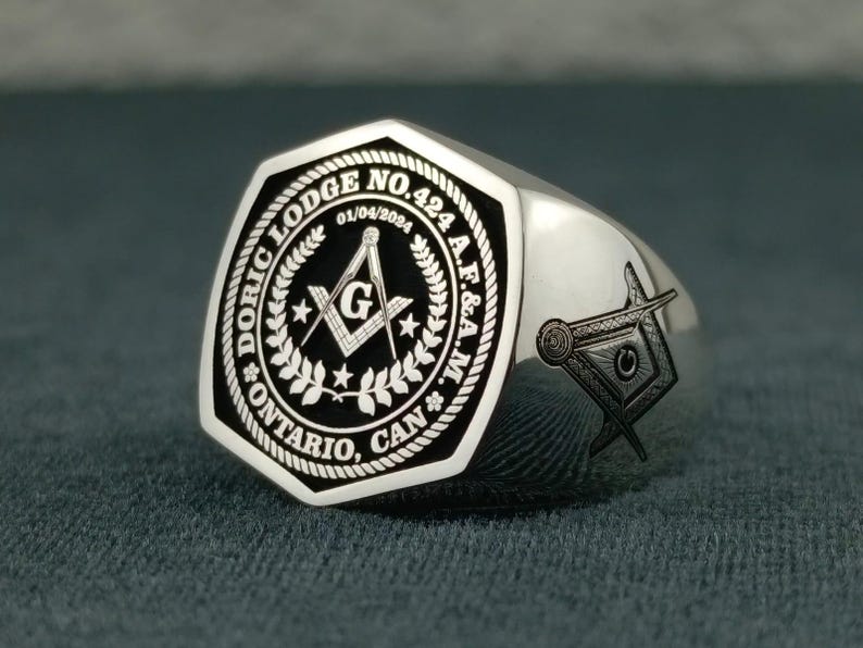 Puede incluir: Anillo mas&oacute;nico de plata con una cara octogonal negra que presenta la inscripci&oacute;n "DORIC LODGE NO. 424 A.F. & A.M. ONTARIO, CAN." y un s&iacute;mbolo mas&oacute;nico. La fecha "01/04/2024" tambi&eacute;n est&aacute; grabada en la cara.