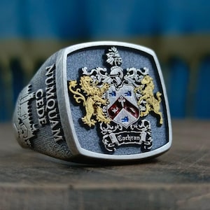 May include: A silver-toned signet ring with a rectangular face. The face features a detailed family crest with gold-colored lions, a shield, and the name "Cochran." The ring's band has the inscription "Numquam Cede" and a castle design.