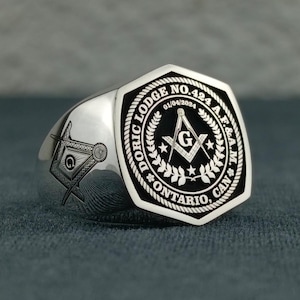 Puede incluir: Un anillo mas&oacute;nico de plata con un dise&ntilde;o grabado en blanco y negro. El anillo presenta el texto "DORIC LODGE NO. 424 A.F.&A.M. ONTARIO, CAN" y la fecha "01/04/2024".