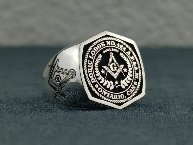 Puede incluir: Un anillo mas&oacute;nico de plata con un dise&ntilde;o grabado en blanco y negro. El anillo presenta el texto "DORIC LODGE NO. 424 A.F.&A.M. ONTARIO, CAN" y la fecha "01/04/2024".