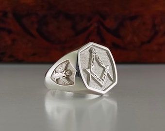 May include: A silver ring with a hexagonal design featuring a detailed Masonic compass and square symbol. The ring also has a smaller design of a winged figure on the side.