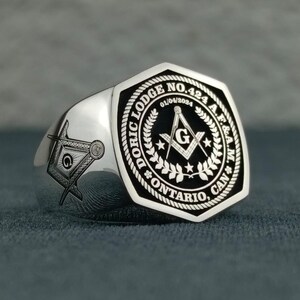 Puede incluir: Un anillo mas&oacute;nico de plata con un dise&ntilde;o grabado en blanco y negro. El anillo presenta una escuadra y un comp&aacute;s, una 'G' y el texto "Doric Lodge No. 424 A.F.&A.M. Ontario, Can."
